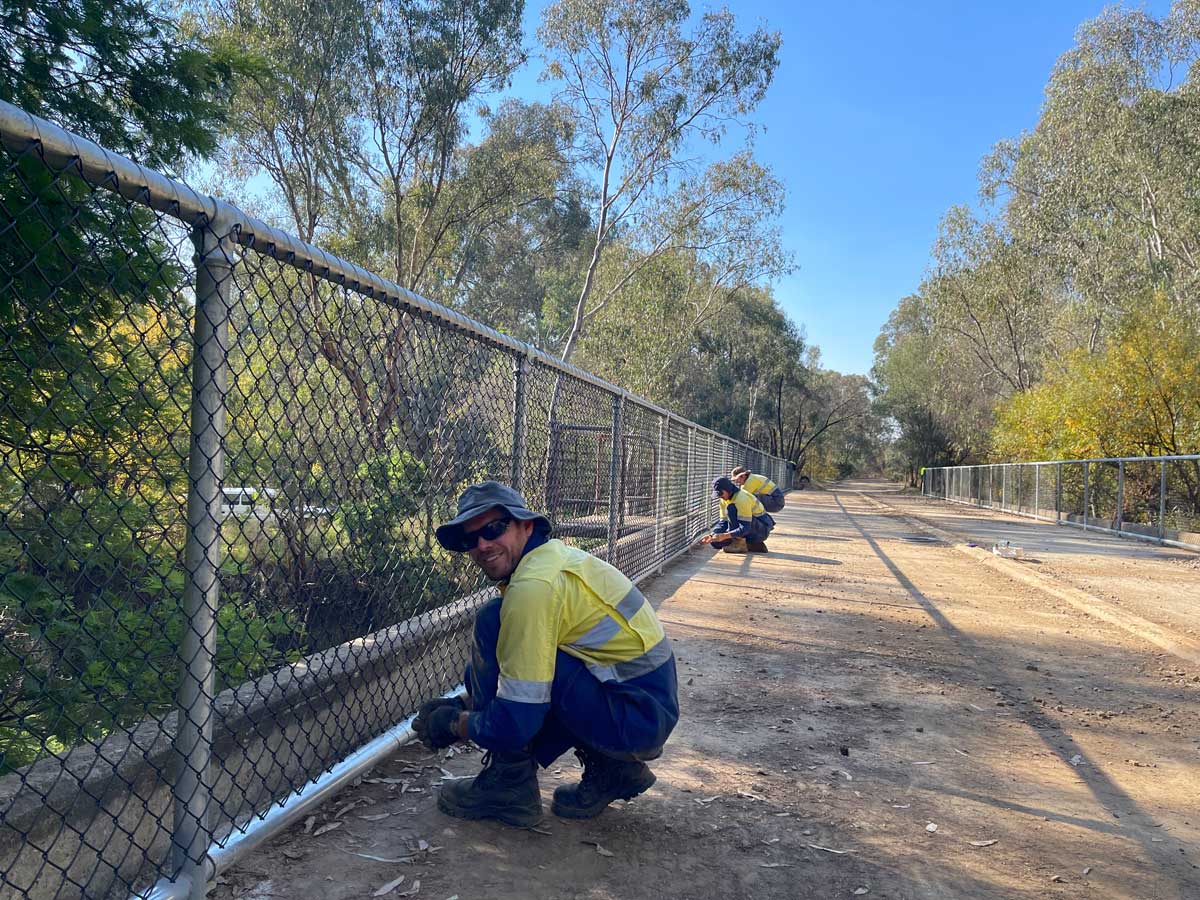 man crouches at base of fence along bridge, working on attaching fence mesh